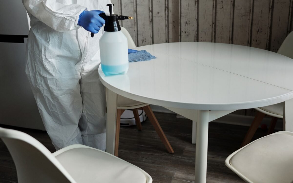 Pessoa em traje de proteção desinfetando uma mesa branca com um pulverizador