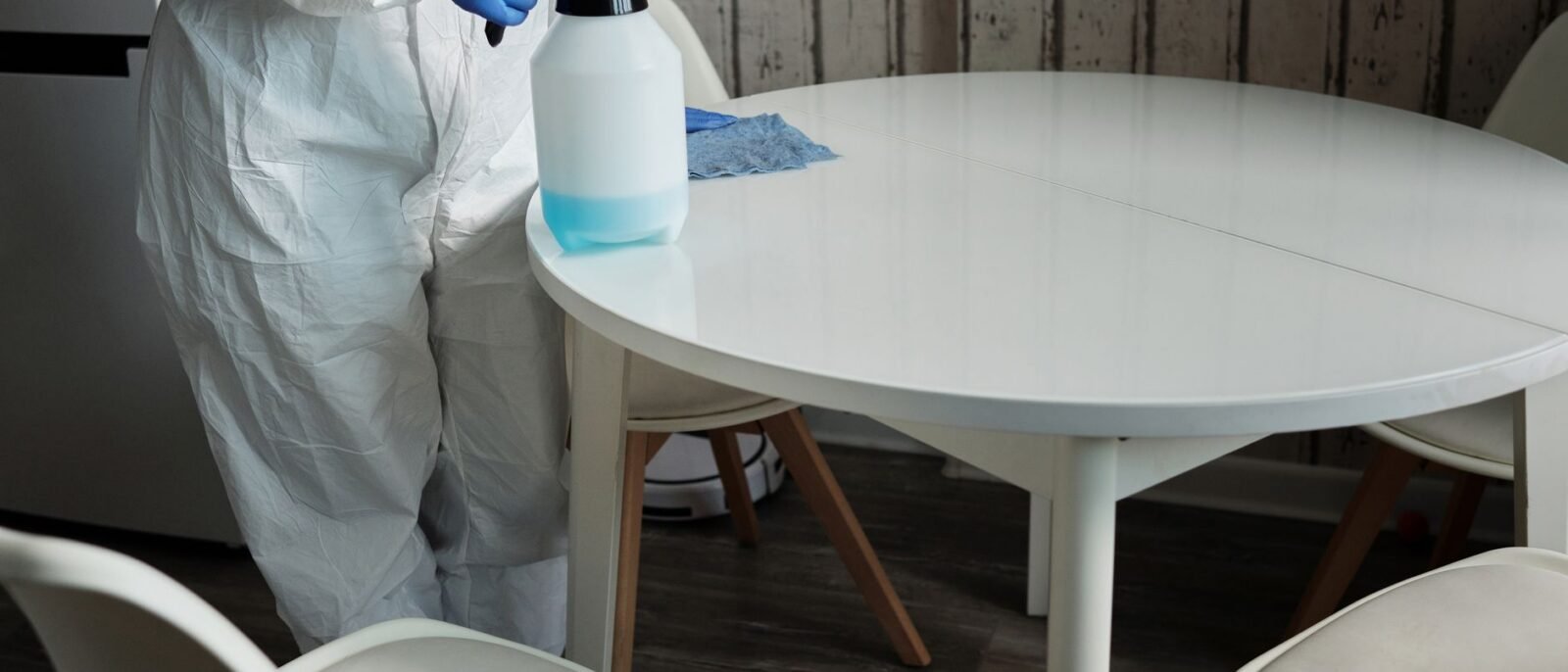 Pessoa em traje de proteção desinfetando uma mesa branca com um pulverizador